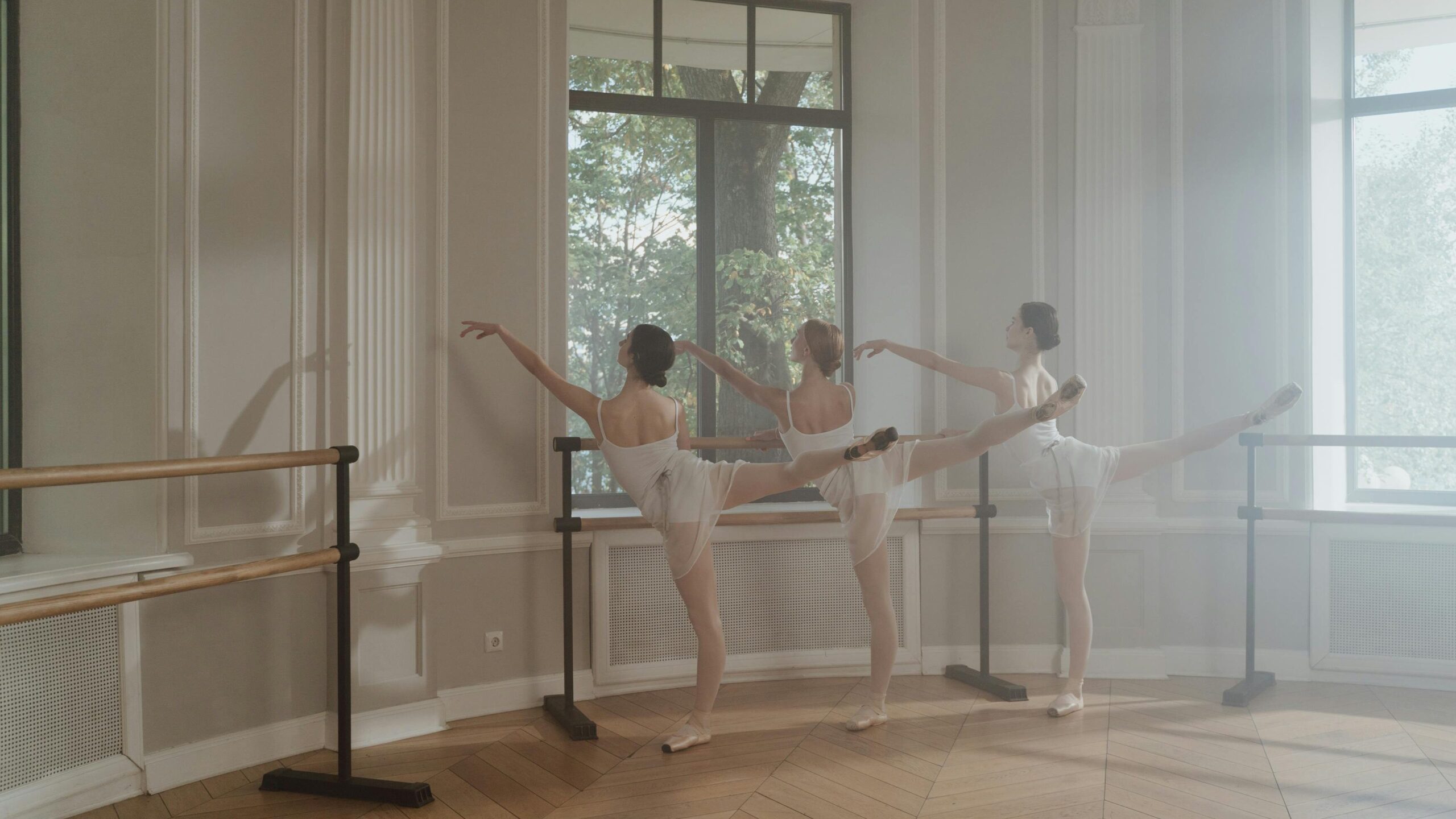 Elegant ballet dancers practicing poses in a sunny dance studio with large windows.