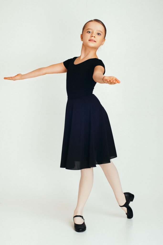 Full length serious girl in black dress dancing with hands apart and looking at camera on white background